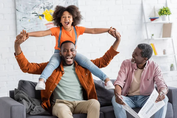 Веселый африканский американский отец и дочь веселятся рядом с дедушкой с газетой дома — стоковое фото