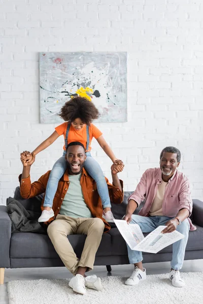 Sonriente afroamericano hombre divirtiéndose con hija cerca de papá sosteniendo periódico en casa - foto de stock