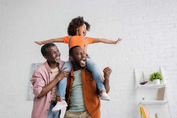 Emocionado hombre afroamericano divirtiéndose con su hija y su padre en casa - foto de stock