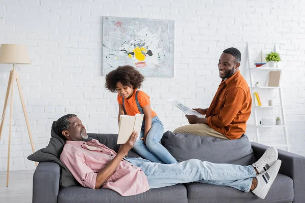 Smiling african american girl sitting near granddad with book and father at home — Stock Photo