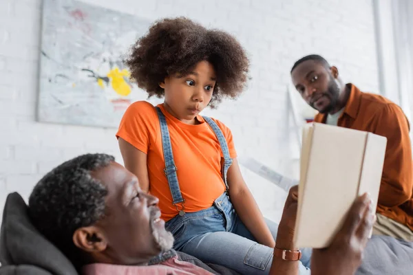 Shocked african american girl looking at book near smiling grandpa and father at home — Stock Photo