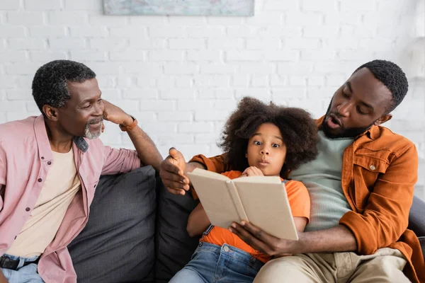 Shocked african american father and daughter reading book near grandparent at home — Stock Photo