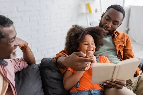 Sonriente hombre afroamericano apuntando a libro cerca de hija y padre borroso en casa - foto de stock