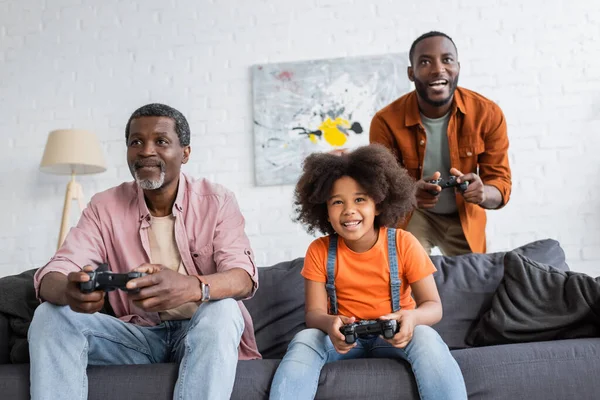 KYIV, UKRAINE - JULY 17, 2021: Cheerful african american kid playing video game with father and granddad at home — Stock Photo