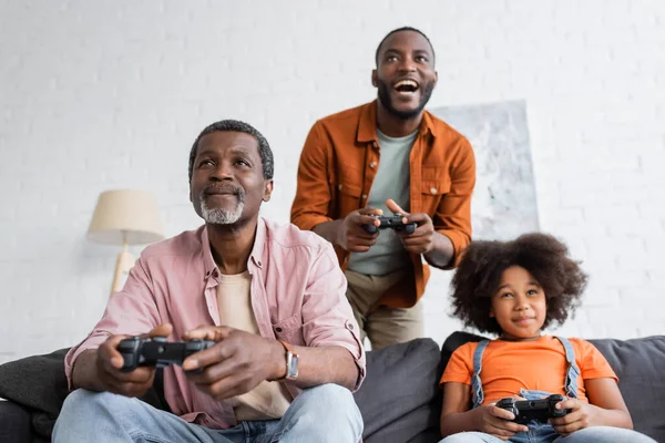 KYIV, UCRANIA - 17 de julio de 2021: El abuelo afroamericano juega un videojuego con su familia en casa - foto de stock