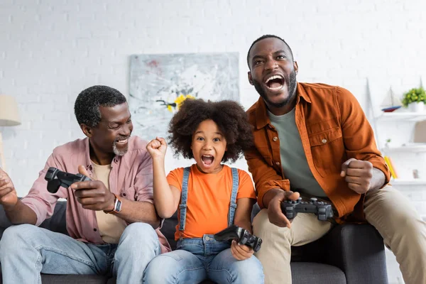 KYIV, UCRAINA - 17 LUGLIO 2021: Ragazza e papà afroamericani eccitati che tengono i joystick vicino al nonno a casa — Foto stock