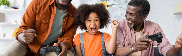 KYIV, UKRAINE - 17 JUILLET 2021 : Enfant afro-américain excité montrant oui geste près des parents avec des joysticks à la maison, bannière — Photo de stock