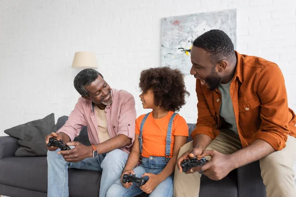 КИЕВ, УКРАИНА - 17 июля 2021 года: Улыбающийся африканский дедушка с бабушкой и бабушкой играют в видеоигры с ребенком и сыном дома — стоковое фото