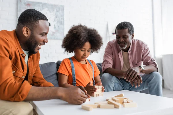 Chica afroamericana jugando bloques de madera juego con papá y abuelo en casa - foto de stock