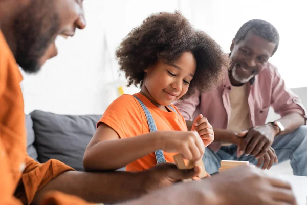Carefree african american girl playing wood blocks game with blurred dad and grandpa at home — Stock Photo