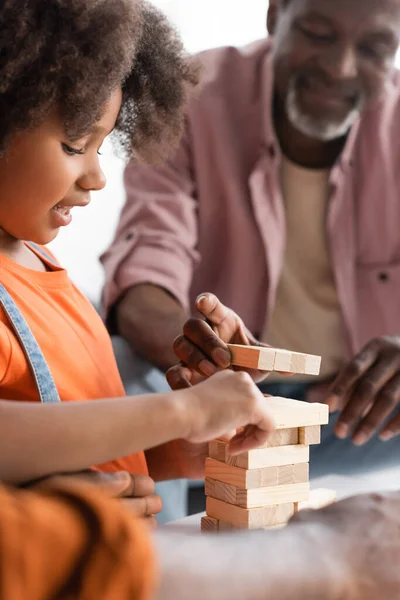 Smiling african american girl playing wood blocks game with blurred dad and grandfather at home — Stock Photo