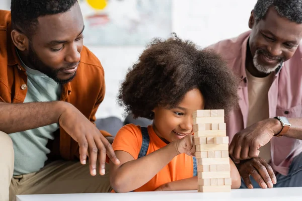 Despreocupado niño afroamericano jugando bloques de madera juego con el padre y el abuelo en casa - foto de stock