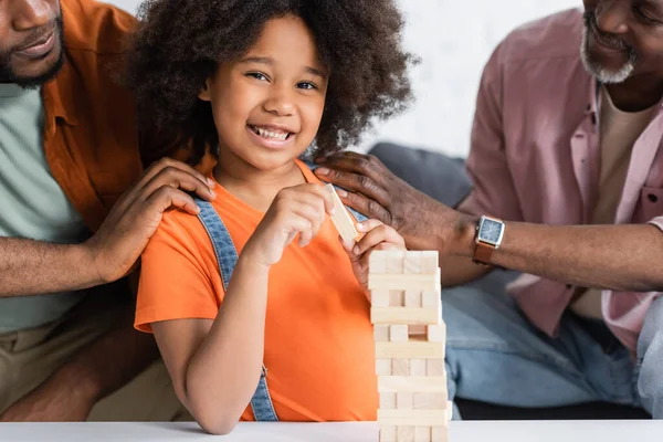 Criança americana africana feliz olhando para a câmera perto de pais e blocos de madeira jogo em casa — Fotografia de Stock