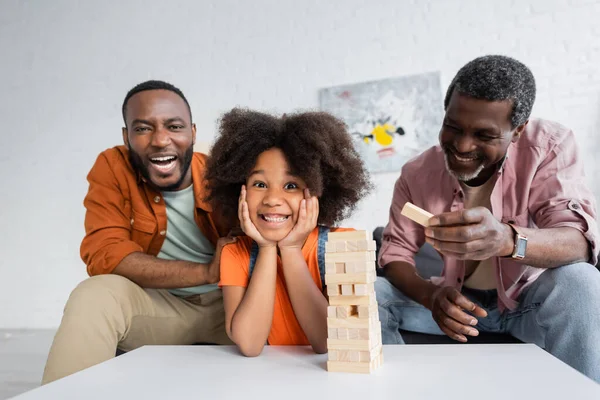 Jovem afro-americano alegre olhando para a câmera perto do avô e do pai enquanto joga jogos de blocos de madeira em casa — Fotografia de Stock