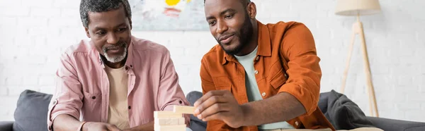 Uomo afroamericano giocare blocchi di legno gioco con papà maturo a casa, banner — Foto stock