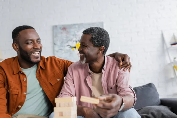 African american man hugging mature father near blurred wood blocks game at home — Stock Photo