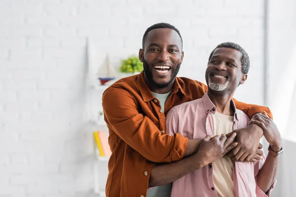 Fröhlich afrikanisch amerikanisch mann umarmt reif papa und schauen auf kamera bei zuhause — Stockfoto