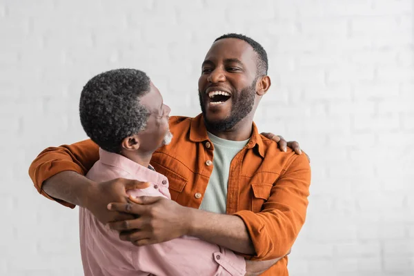 Positivo afroamericano hijo abrazando maduro padre en sala de estar - foto de stock