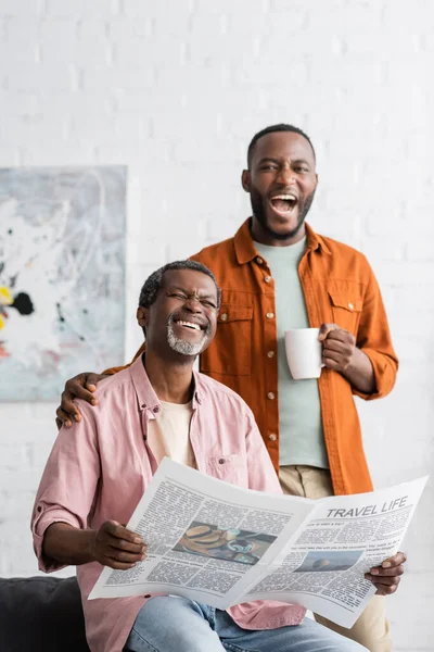 Веселий афроамериканець тримає в руках чашку кави біля тата з газетою вдома. — стокове фото