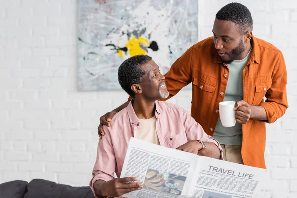 African american man holding cup of coffee and talking to father with newspaper at home — Stock Photo