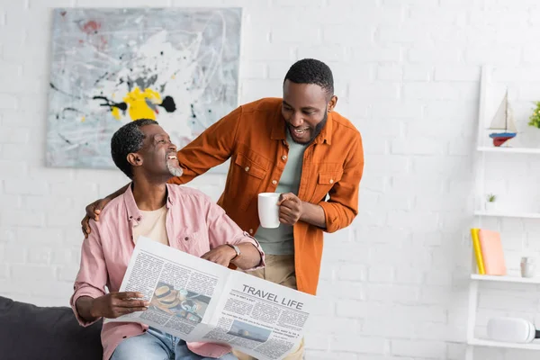 Smiling african american man holding cup and hugging father with newspaper in living room — Stock Photo