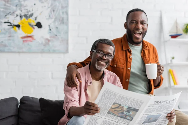 Uomo afroamericano spensierato con coppa abbracciando padre maturo con giornale in soggiorno — Foto stock