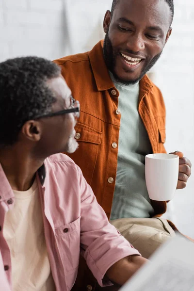 Un homme afro-américain positif tenant une tasse de café près d'un père flou avec un journal à la maison — Photo de stock
