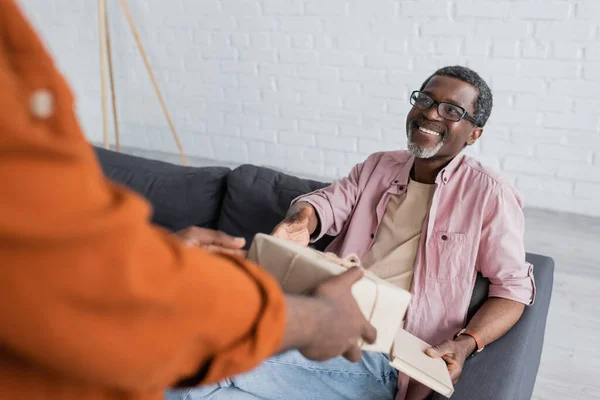 Cheerful african american man with book taking gift box from son during father day celebration at home — Stock Photo