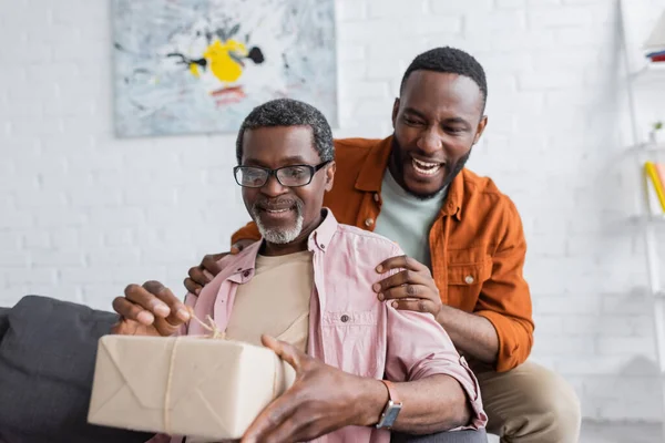 Sorrindo afro-americano homem abraçando o pai com presente durante o dia do pai em casa — Fotografia de Stock