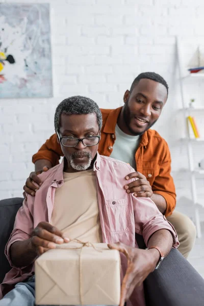 African american man hugging shocked parent with gift during father day celebration at home — Stock Photo