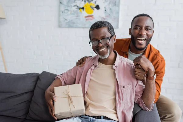 Веселий афроамериканський чоловік середнього віку тримає подарунок біля сина під час святкування дня батька вдома — стокове фото