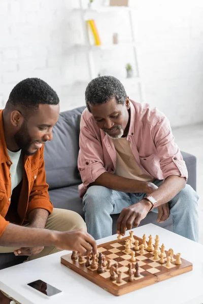 Hombre afroamericano jugando ajedrez con hijo sonriente cerca de teléfono inteligente en casa - foto de stock