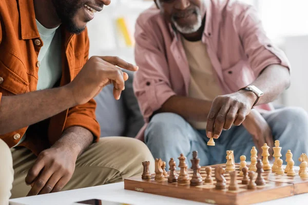 Розбитий вигляд усміхненого афро - американського чоловіка, який вказує на шахову дошку, граючи вдома з татом. — стокове фото