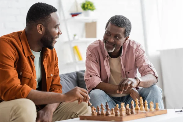 Улыбающийся африканский американец играет в шахматы со взрослым сыном в гостиной — стоковое фото