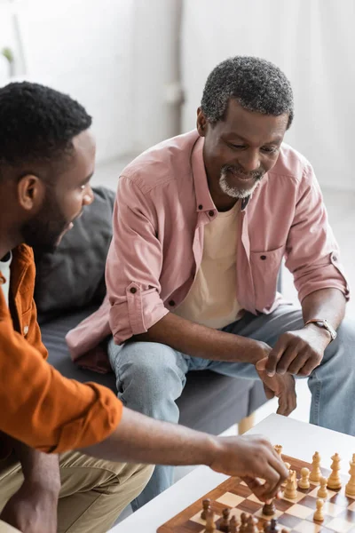 Mature african american dad looking at chess board while playing with blurred son at home — Stock Photo