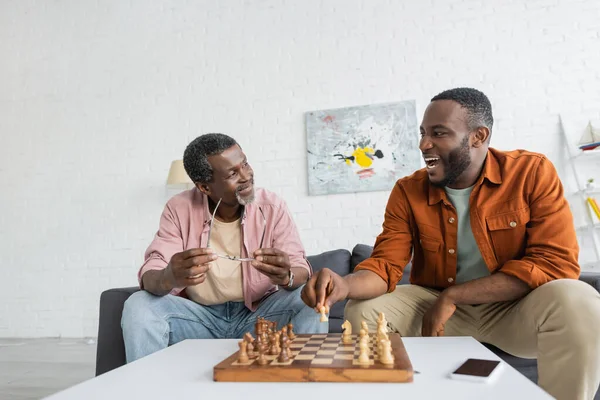 Overjoyed african american man playing chess with mature dad near smartphone with blank screen at home — Stock Photo