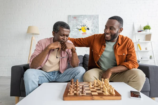 Sonriente hombre afroamericano abrazando a papá enfocado mientras juega al ajedrez en la sala de estar - foto de stock