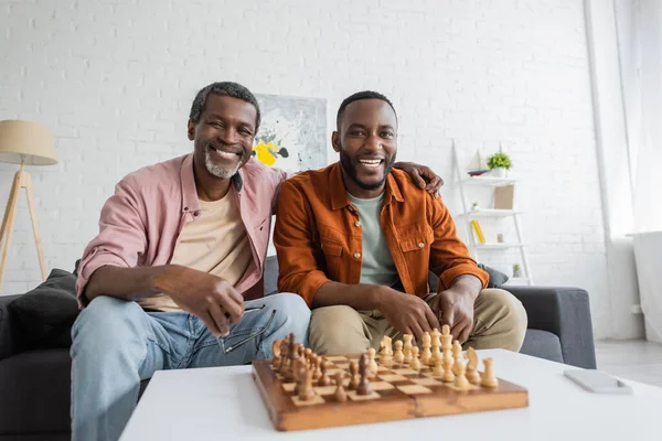 Sonriente hombre afroamericano con anteojos abrazando hijo y mirando a la cámara cerca de ajedrez y teléfono inteligente en casa - foto de stock