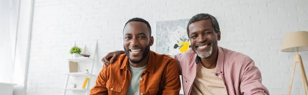 Positive afrikanisch-amerikanische Mann umarmt erwachsenen Sohn und schaut in die Kamera zu Hause, Banner — Stockfoto