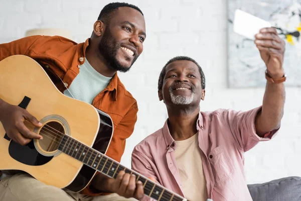 Smiling african american father taking selfie with son playing acoustic guitar at home — Stock Photo