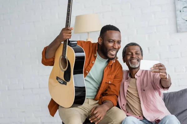 Positivo afroamericano padre e hijo con guitarra acústica tomando selfie en casa - foto de stock