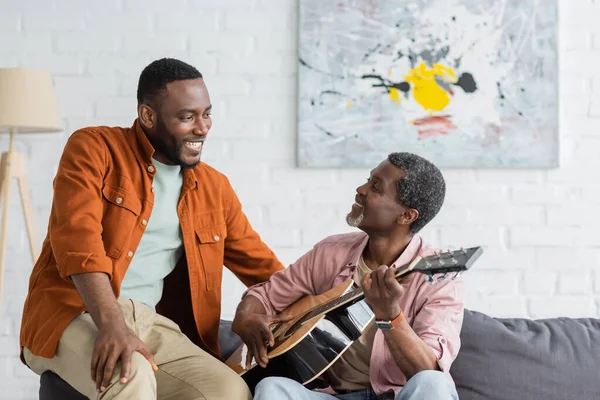 Зрелый африканский отец-американец играет на акустической гитаре рядом с сыном на диване дома — стоковое фото