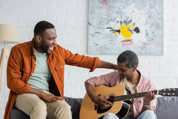 Fröhlich afrikanisch-amerikanischer Mann umarmt reifen Vater, der zu Hause Akustikgitarre spielt — Stockfoto