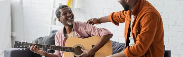 Alegre hombre afroamericano tocando la guitarra acústica cerca de hijo en casa, pancarta - foto de stock
