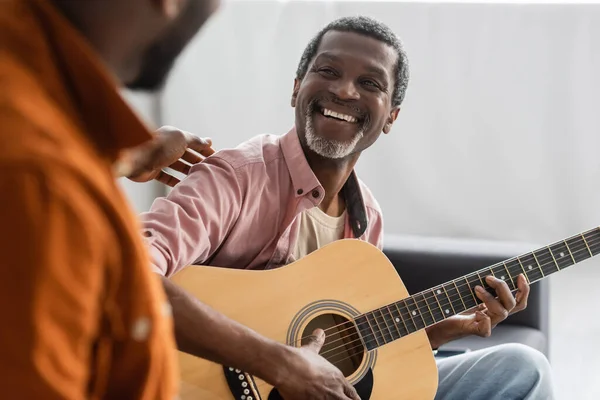 Щасливий батько-афроамериканець, який грає на акустичній гітарі біля розмитого сина вдома. — стокове фото