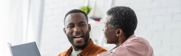 Веселий афроамериканець розмовляє з батьком біля фотоальбому вдома, банер. — стокове фото