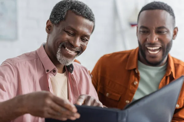 Carefree african american man pointing at blurred photo album near son at home — Stock Photo