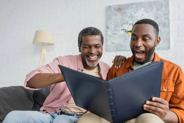 Alegre y emocionado afroamericano padre e hijo mirando álbum de fotos en casa - foto de stock