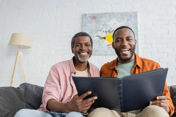 Sorrindo afro-americano pai e filho segurando álbum de fotos e olhando para a câmera em casa — Fotografia de Stock