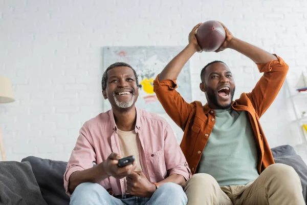 Cheerful african american man holding remote controller near son with rugby ball at home — Stock Photo